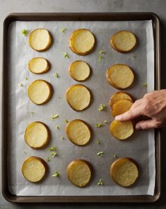 A side view of the baking sheet in the oven with golden, slightly crispy potato rounds visible through the oven door. The oven light is on for a warm, appetizing effect.