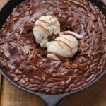 ustic-style dessert presented in a round, black cast iron skillet. The dessert is a rich, chocolate brownie topped with two scoops of vanilla ice cream.
